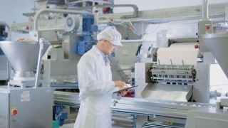 两个年轻的食品工厂员工讨论与工作有关的事情。男性技术员或质量经理使用平板电脑工作。他们戴着白色的卫生帽和工作袍。